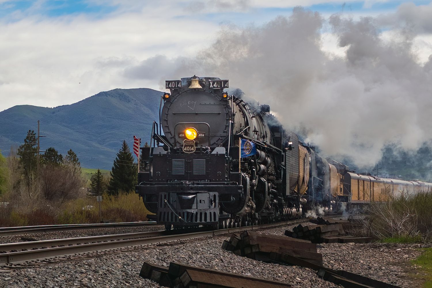 A vintage steam locomotive billows smoke as it travels through a scenic mountain valley on a clear day.