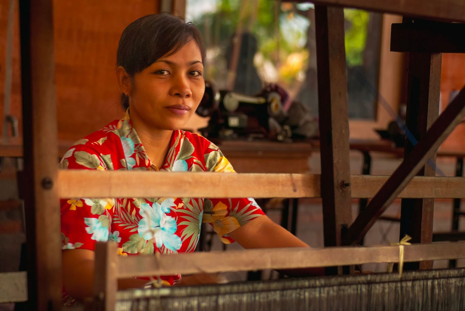 Close-up portrait of a woman in a floral shirt sitting at a traditional wooden weaving loom indoors.