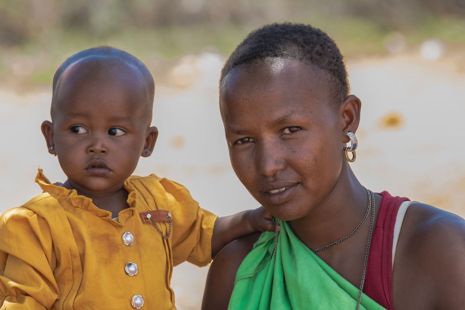 African mother in green dress holding young baby in yellow outfit in rural setting.