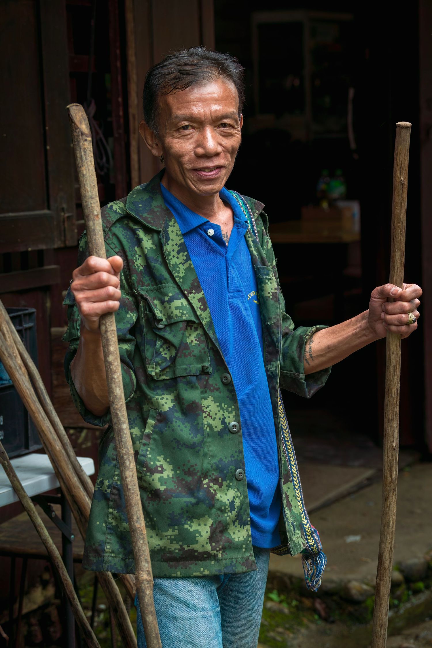 An elderly Asian man in a camouflage jacket smiles while holding two wooden walking sticks outdoors.
