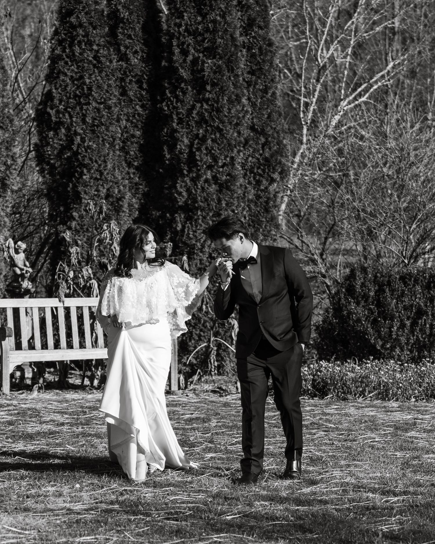 Black and white photo of bride and groom walking in garden, groom kissing brides hand near wooden bench.