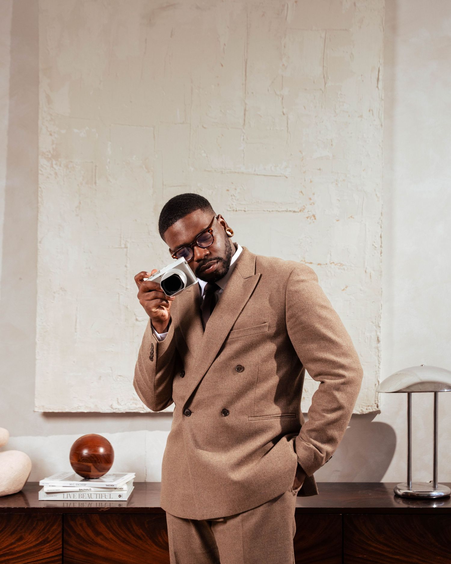 Stylish man in camel suit holds camera to eye while posing in front of textured white wall.