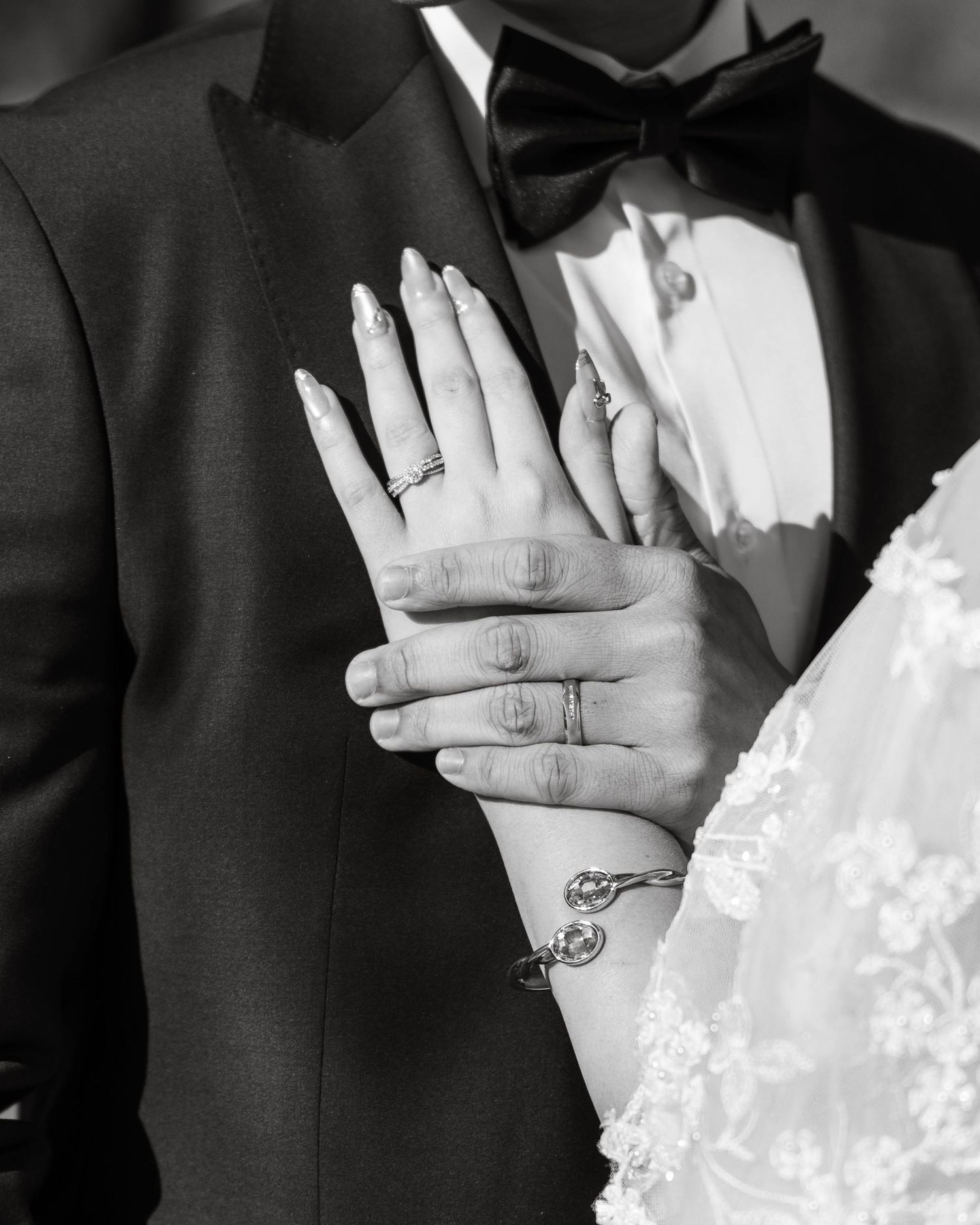 Black and white close-up of bride and groom hands showing wedding rings, with bride wearing lace dress and bracelet.