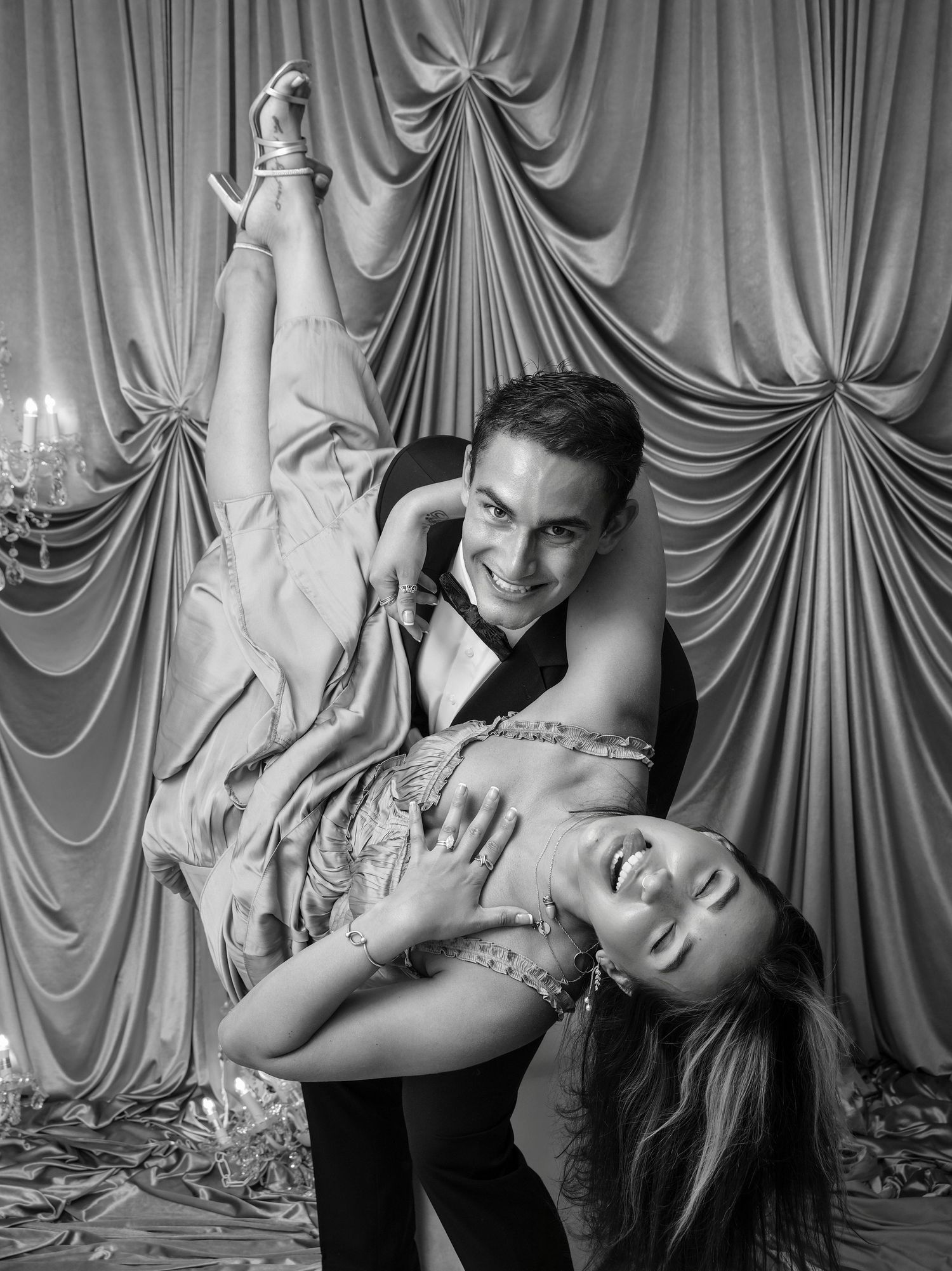 Black and white photo of smiling man dipping woman in glamorous gown before draped curtain backdrop.