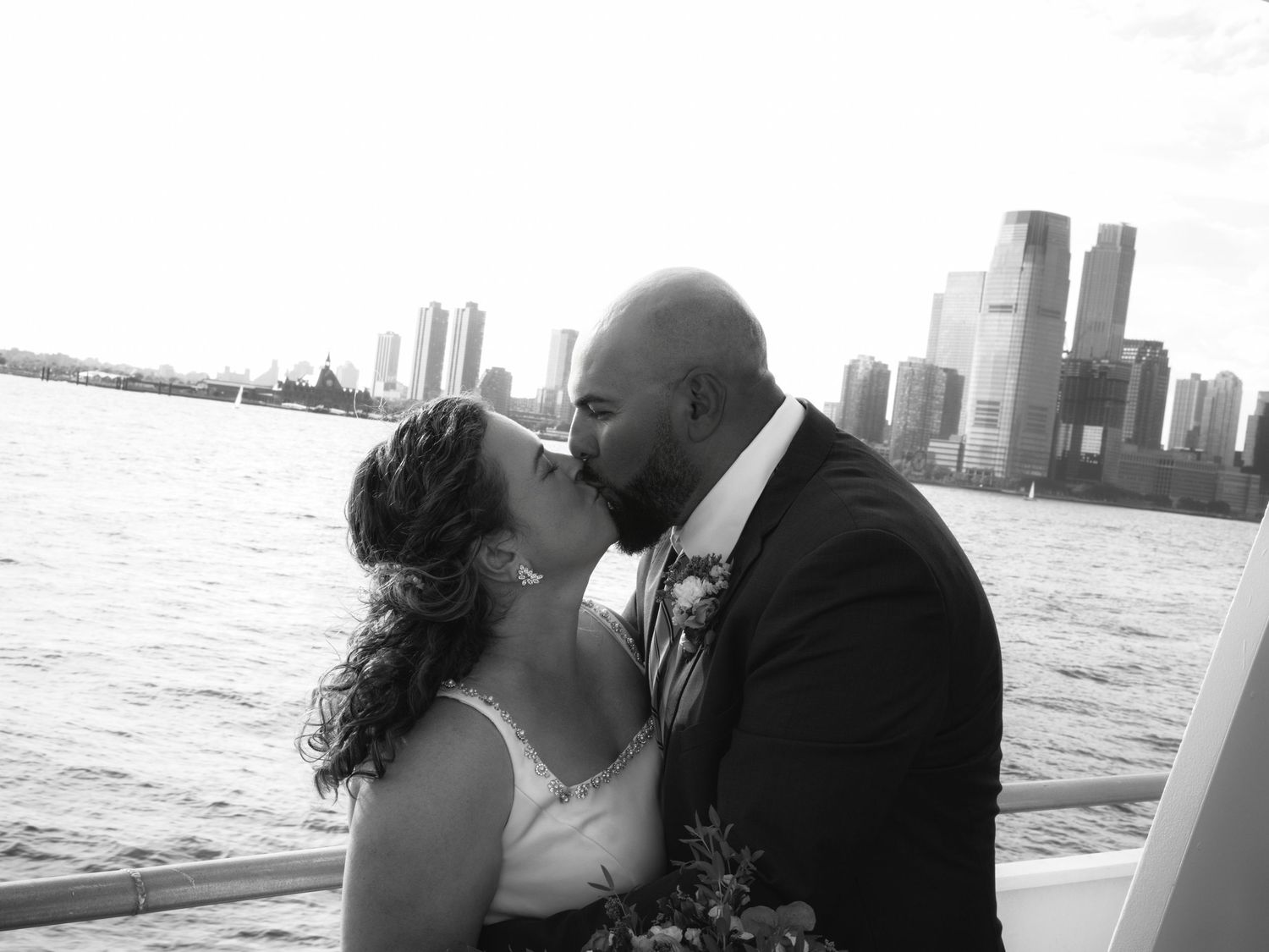 Black and white photo of bride and groom kissing on boat with city skyline across the water.