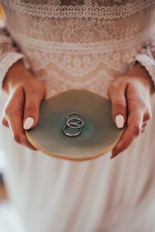Bride holding ceramic dish with two silver wedding rings in lace dress.