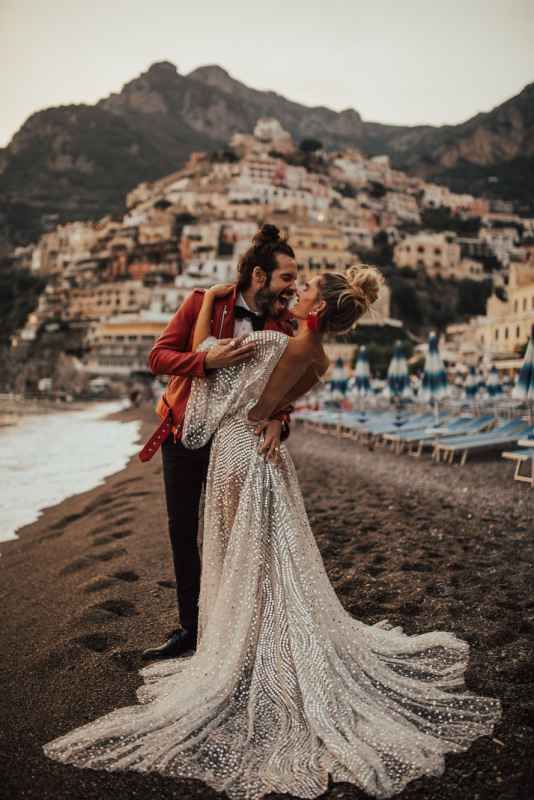 Bride and groom kissing on a black sand beach with the colorful cliffs of Positano behind them.