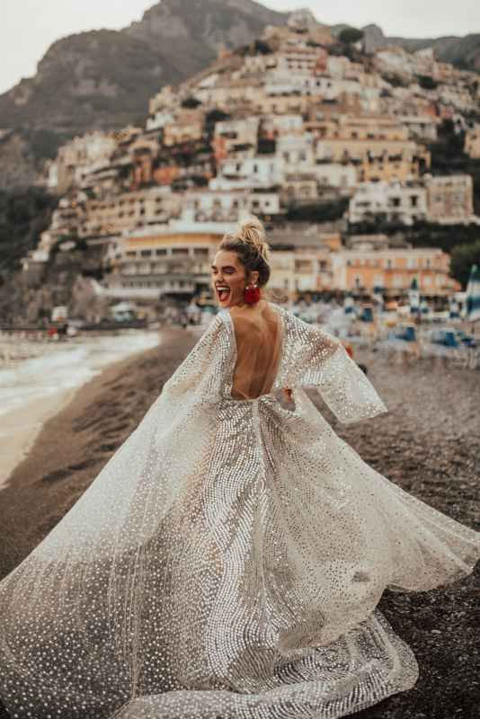 Bride twirling in stunning backless sequin gown on beach with Positano Italy hillside behind.
