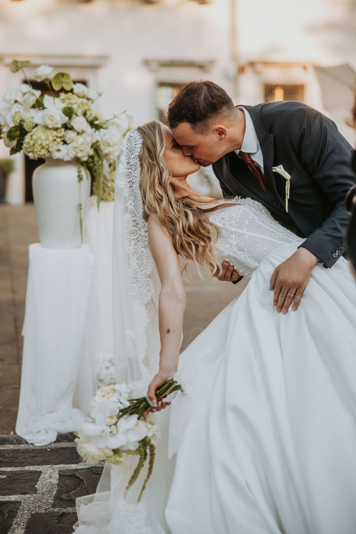 Groom dips bride for a romantic kiss beside white floral arrangements at outdoor wedding ceremony.