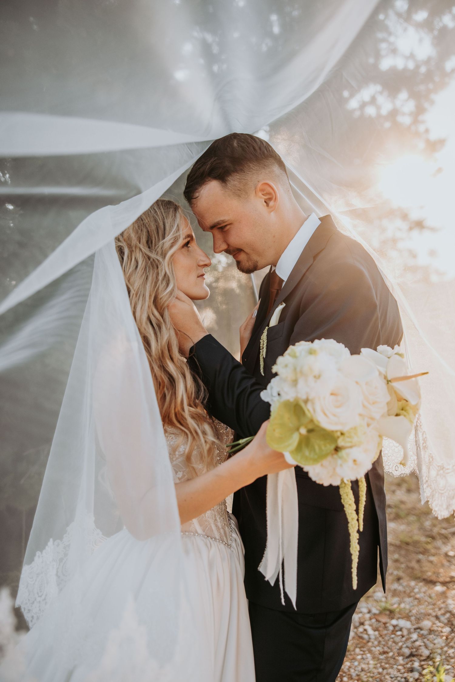 Bride and groom share romantic moment under flowing veil with white bouquet in golden light.
