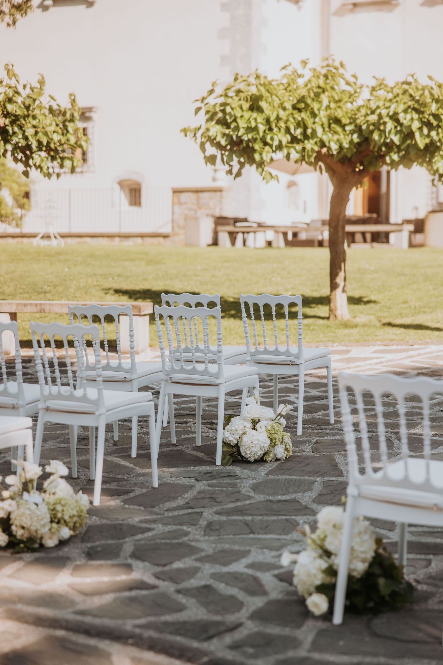 White chairs arranged for outdoor wedding ceremony on stone patio beside green lawn.