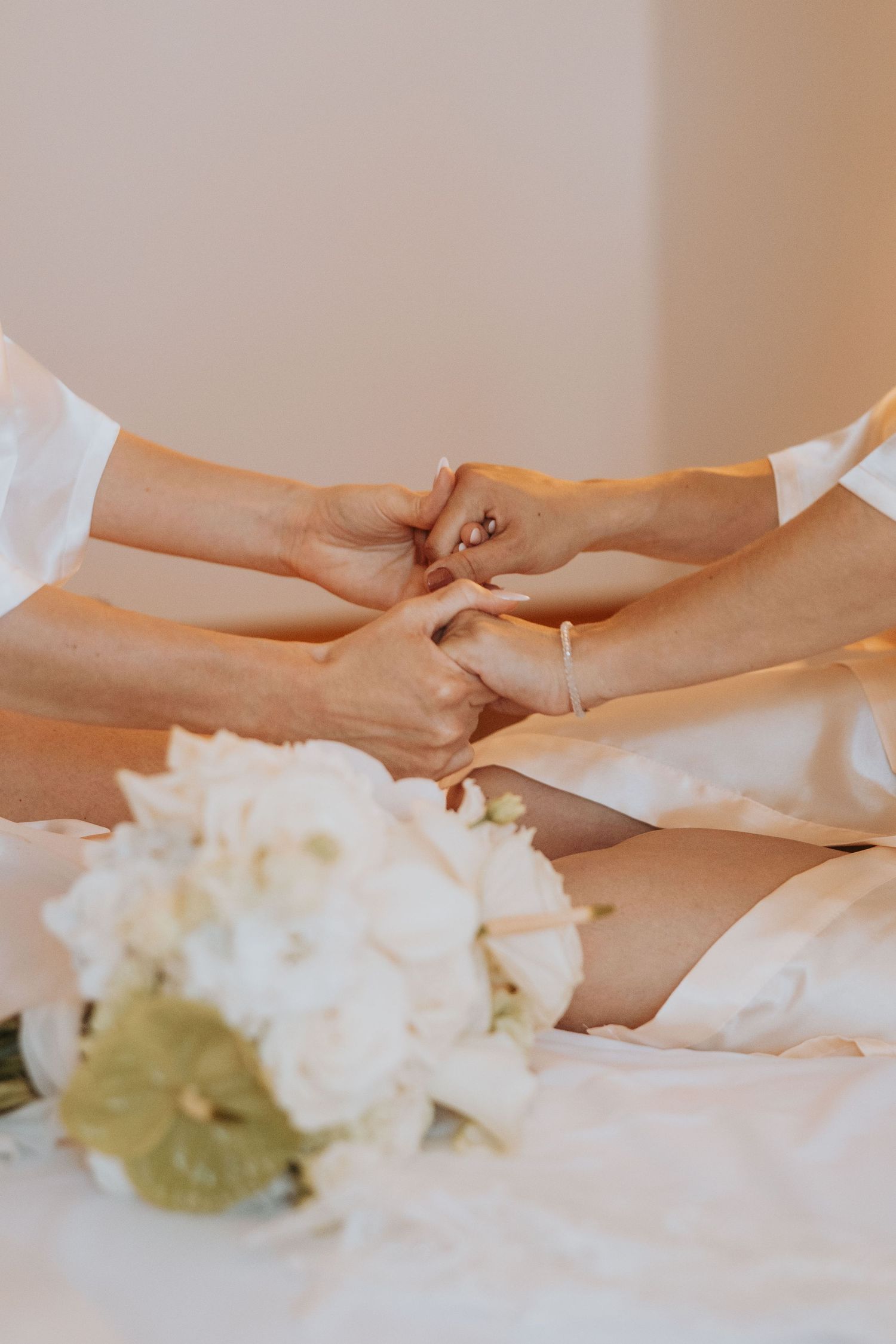 Bride and bridesmaid in white robes holding hands with bridal bouquet of white flowers in foreground.