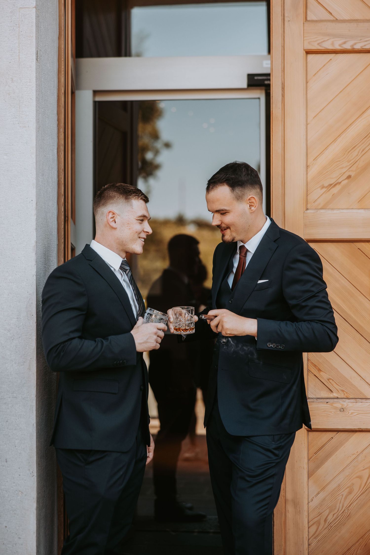 Two men in dark suits toasting with glasses outside wooden doors at a formal event.