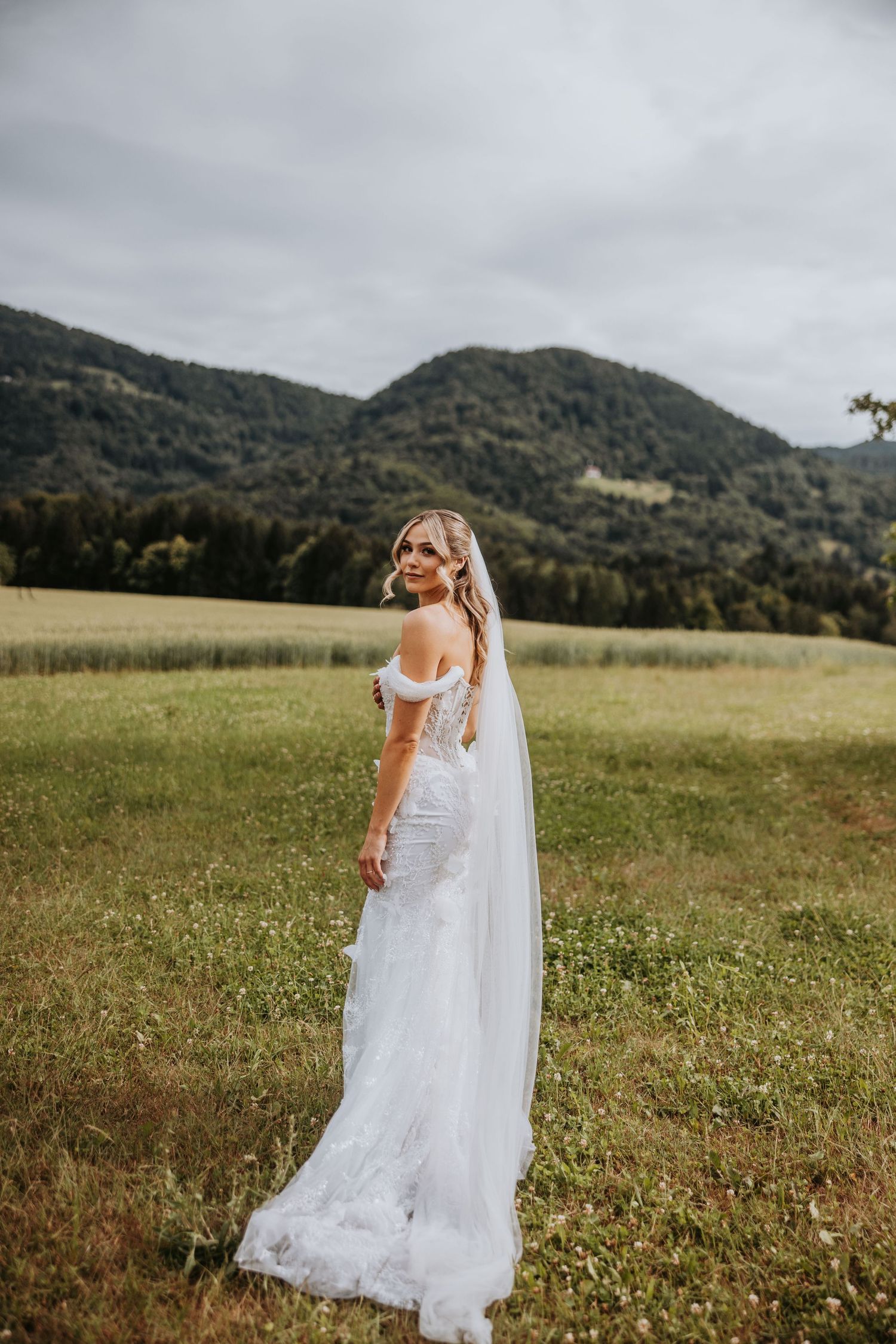 Bride in lace off-shoulder wedding gown with cathedral veil posing in green meadow with mountain backdrop.
