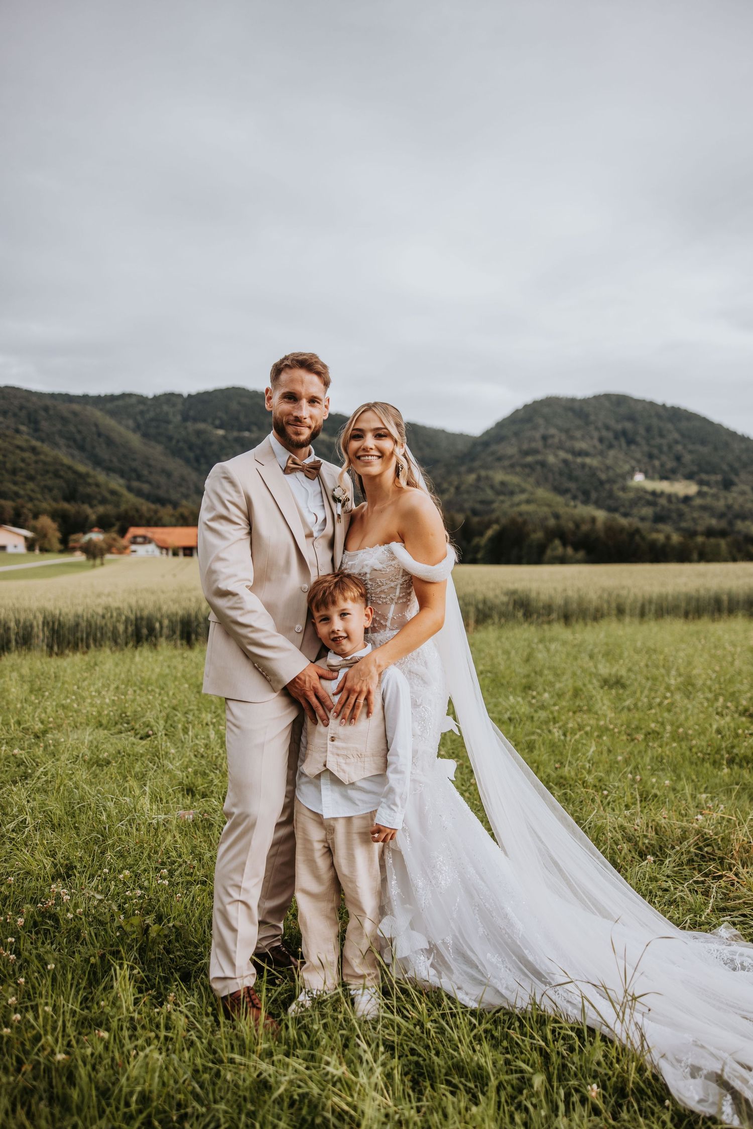 Bride and groom in beige suit pose with young child in green meadow with mountain backdrop on wedding day.