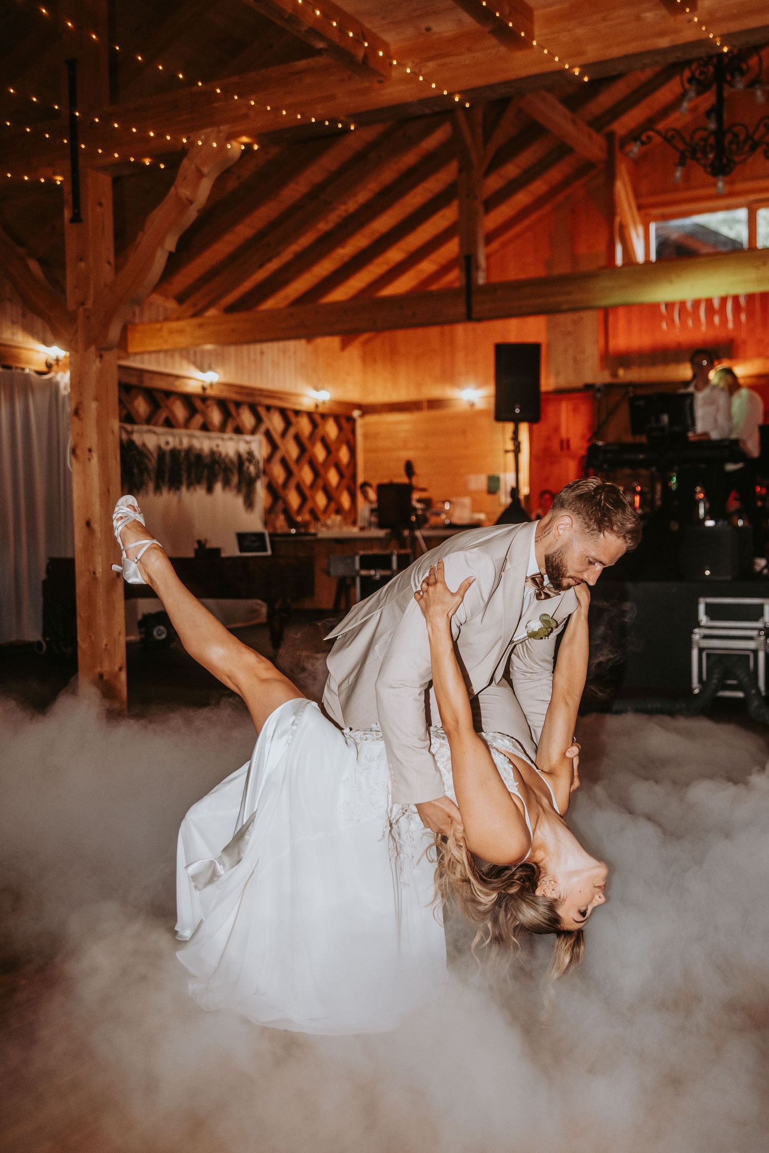 Bride and groom perform dramatic first dance dip surrounded by atmospheric fog in rustic barn venue.