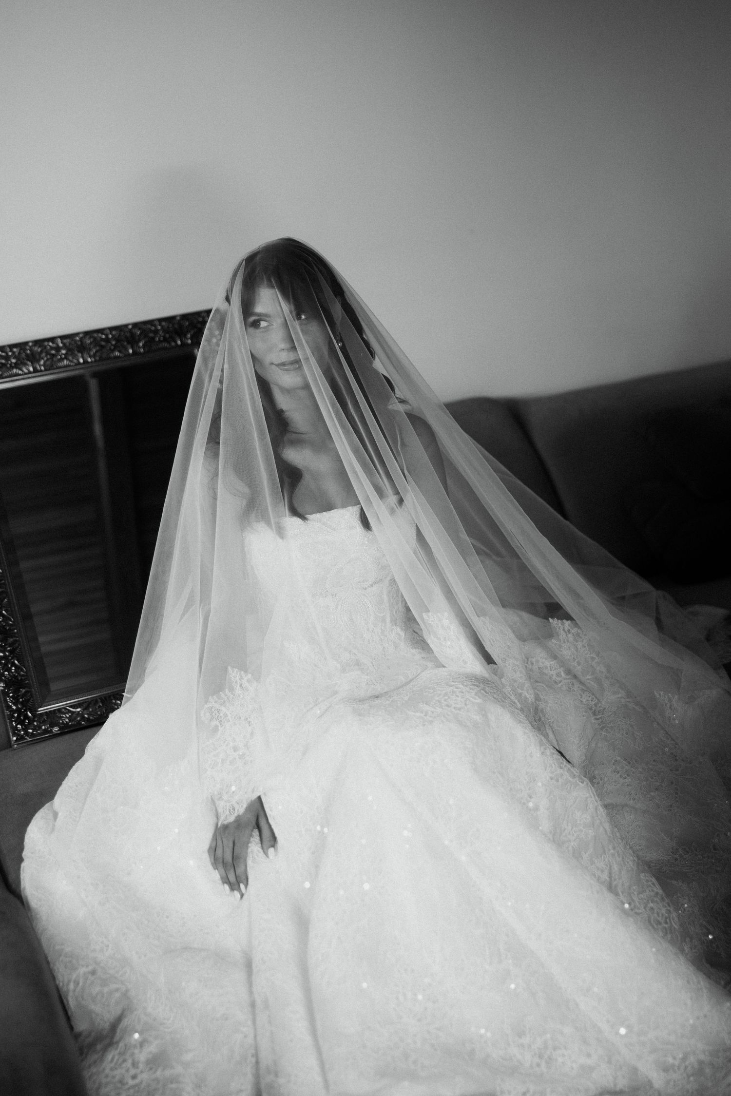 Black and white photo of a bride in a lace wedding gown with a long veil draped over her face, seated elegantly.