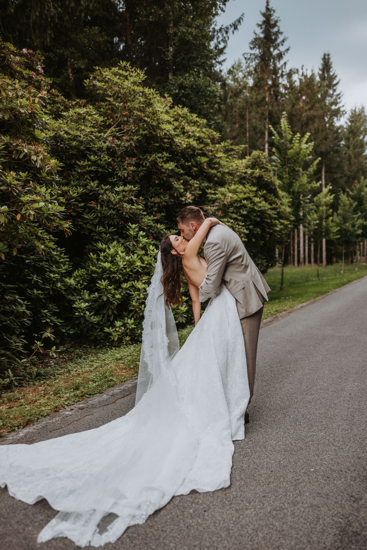 A groom dips his bride for a romantic kiss on a tree-lined path, her white gown and veil trailing elegantly.
