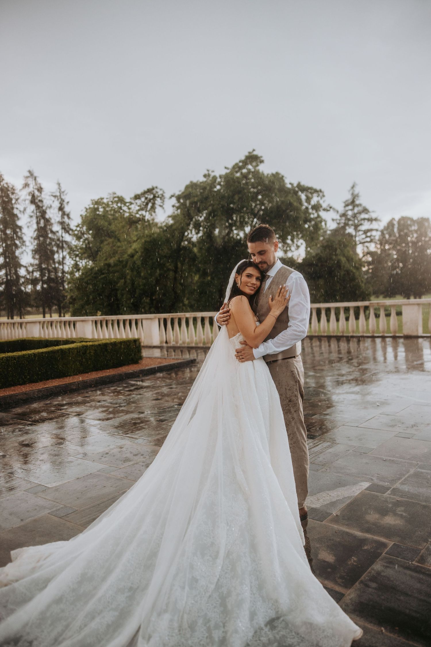 Bride and groom embracing romantically on rain-soaked terrace with lush garden in background.