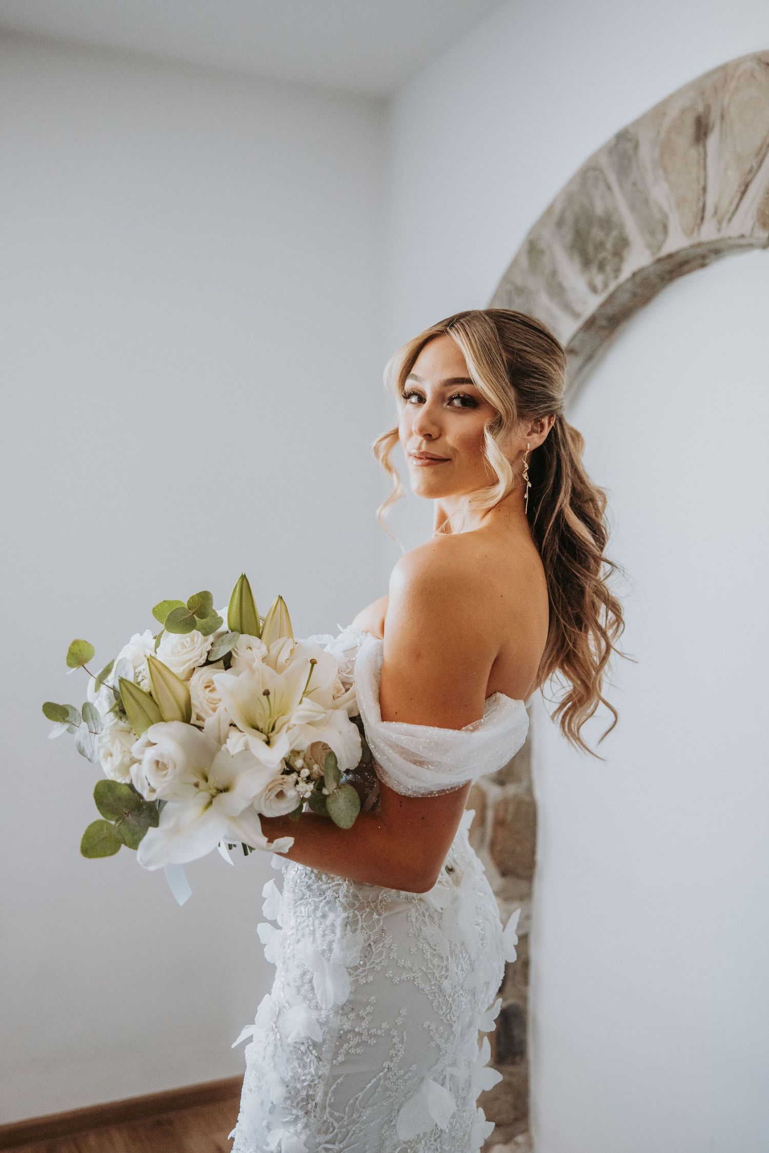 Bride in off-shoulder lace gown holding white lily and rose bouquet, smiling beside stone arch.