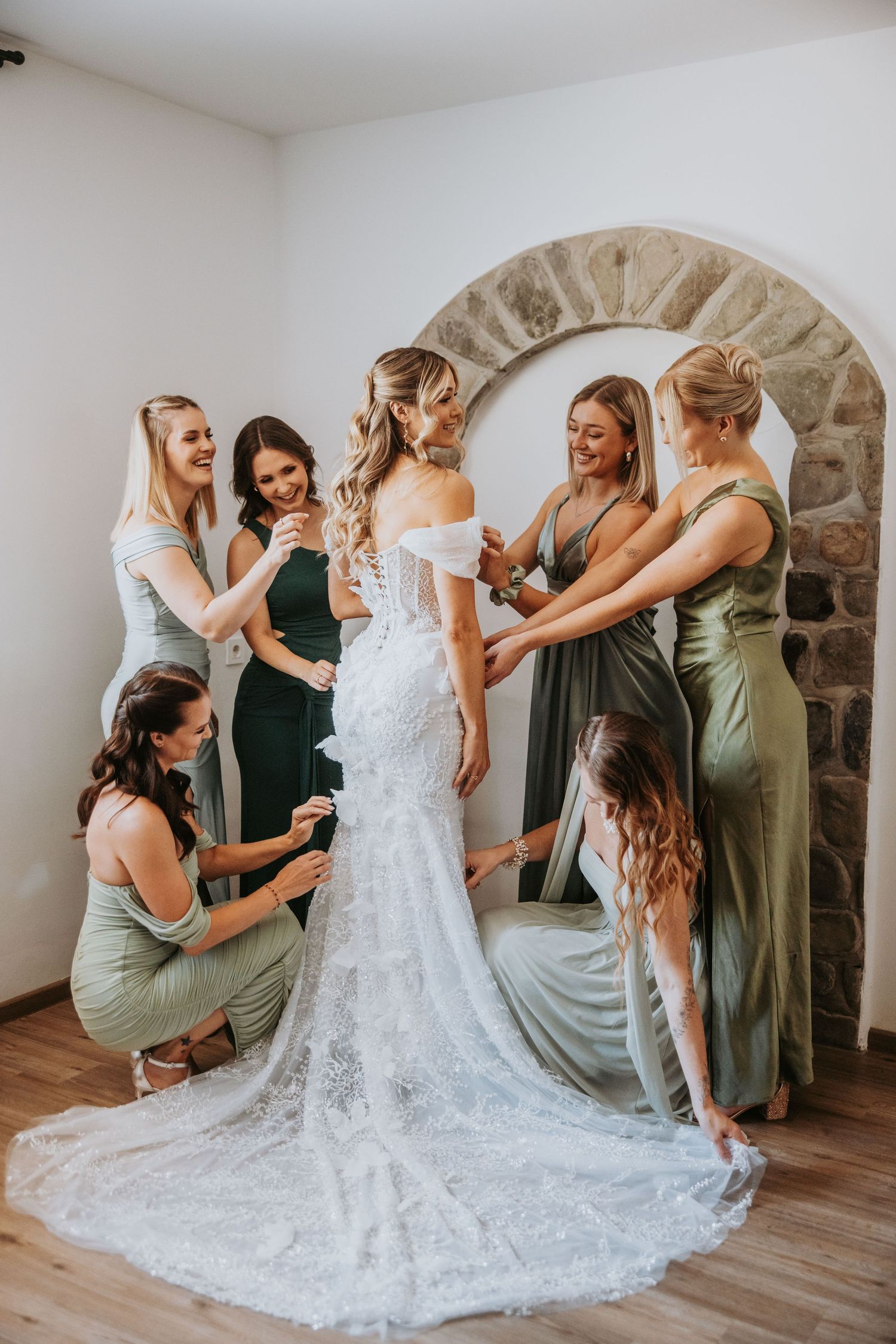 Bridesmaids in sage green dresses help bride into white lace wedding gown near stone arch mirror.