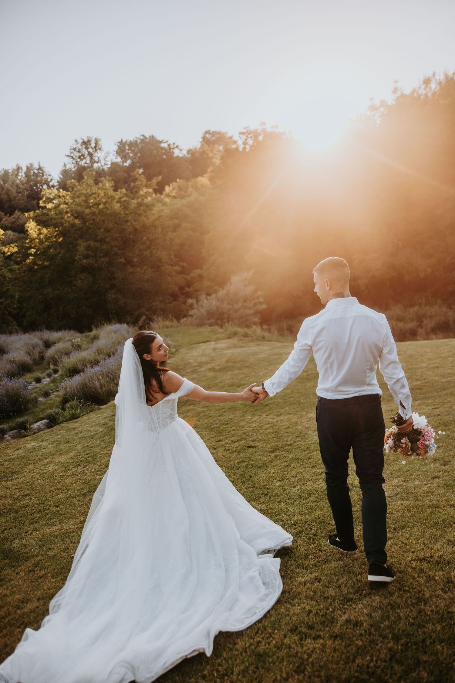 Bride and groom walk hand in hand through a sunlit garden, her long veil trailing behind.