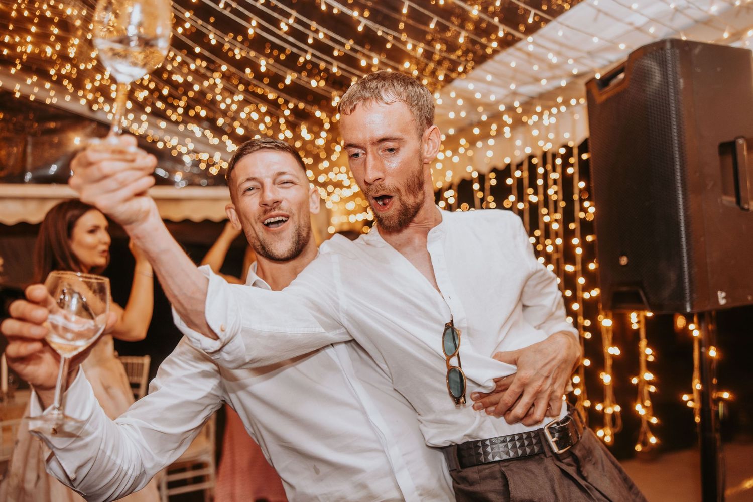 Two men celebrating at wedding reception under warm fairy lights with drinks.