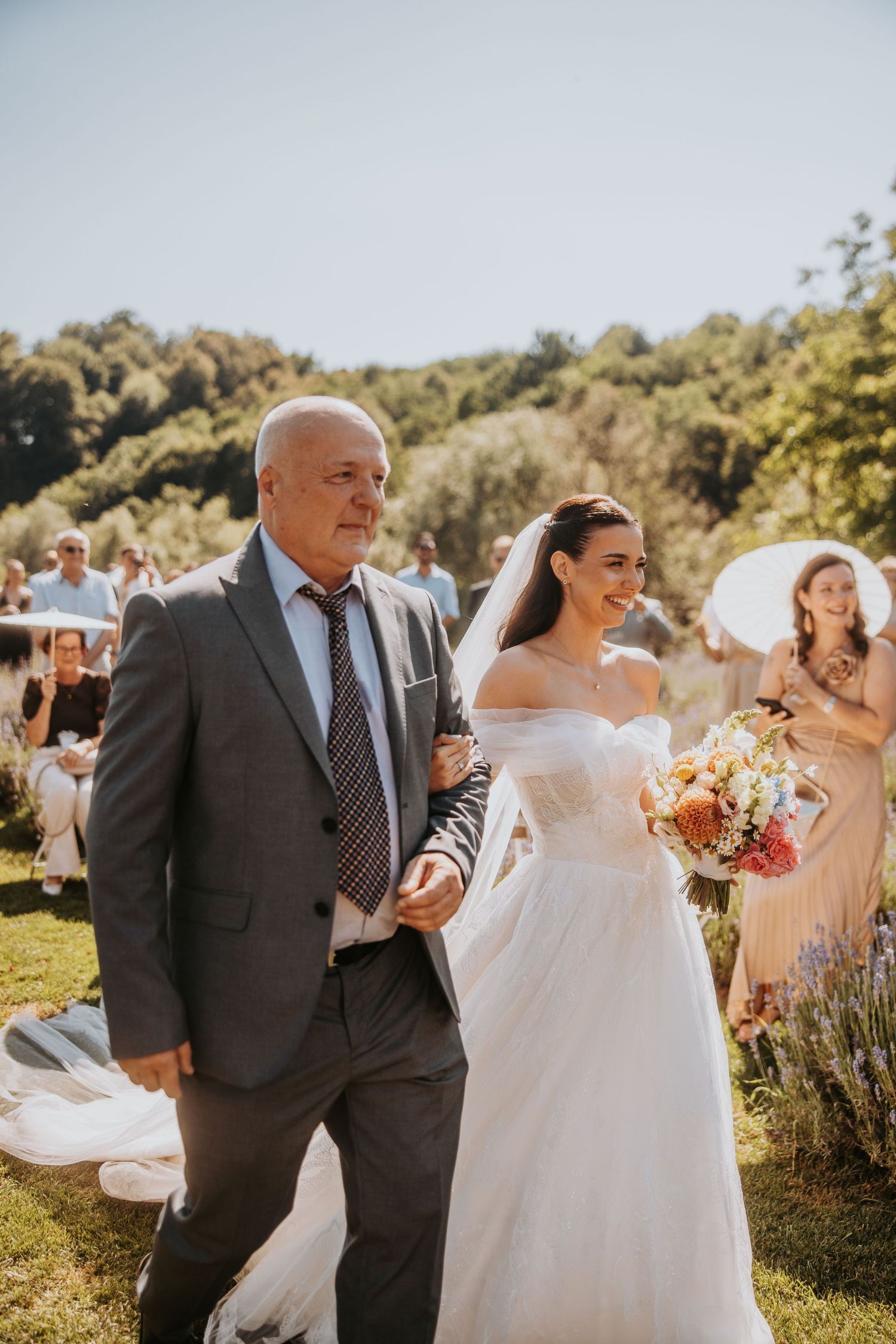 Bride in an off-shoulder wedding gown holding a colorful bouquet walks down the aisle with her father.
