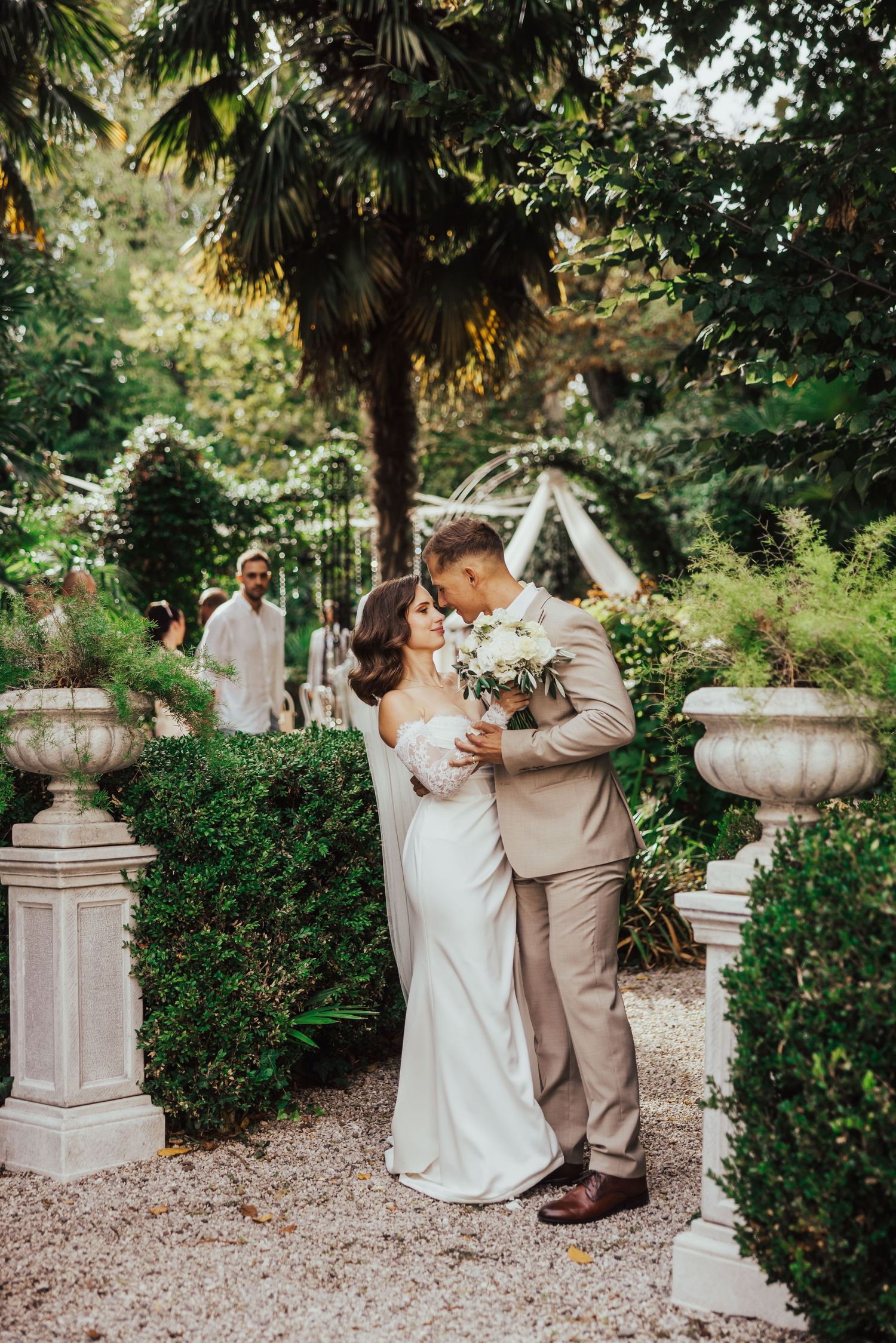 Bride and groom sharing a romantic kiss in a lush garden with stone urns and palm trees.