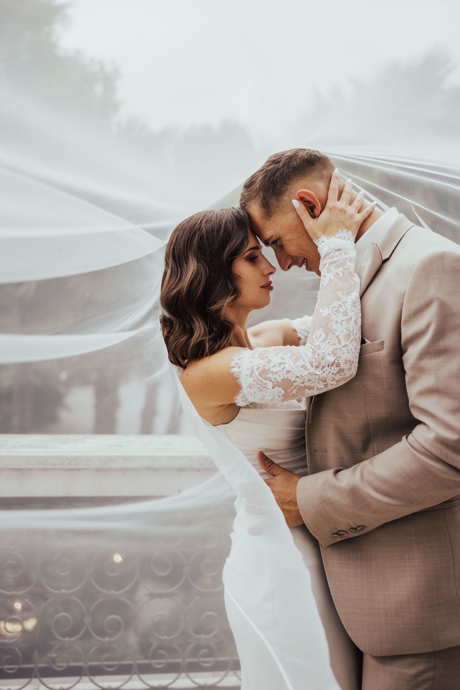 Romantic wedding couple sharing an intimate moment under a flowing white veil, bride in lace gown and groom in beige suit.
