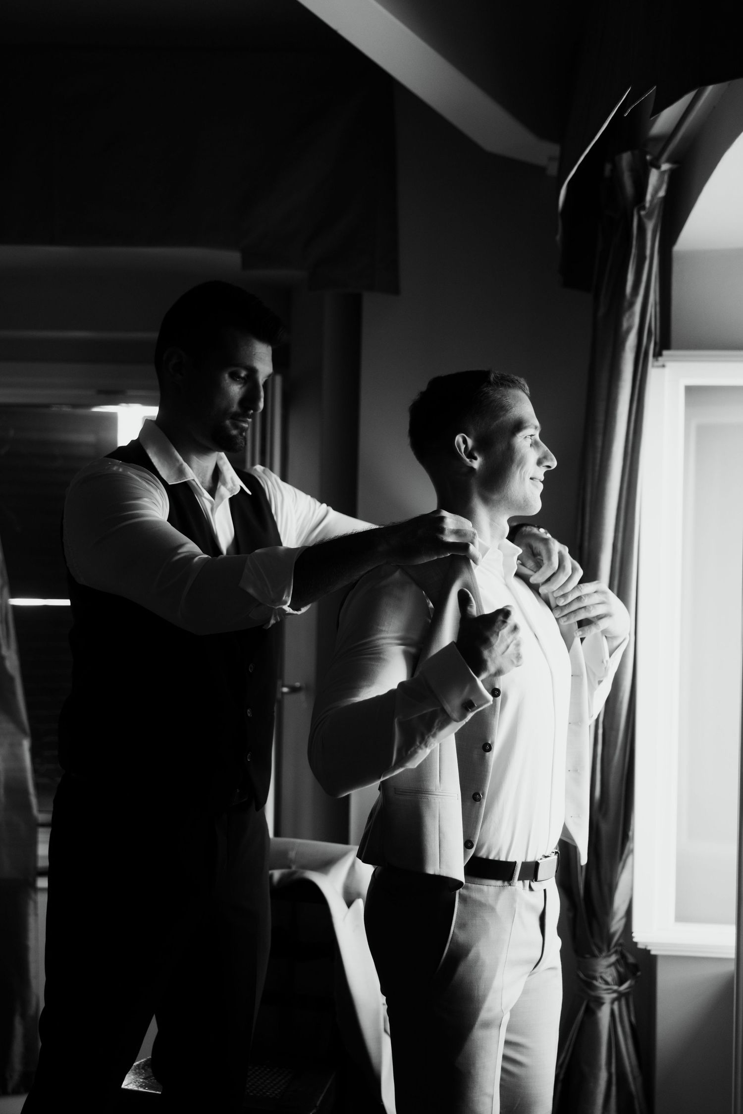 Black and white photo of a man helping a groom put on his suit jacket before a wedding ceremony.
