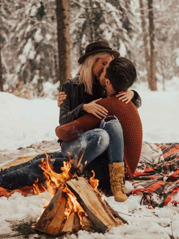 Romantic couple sharing a kiss by a campfire in a snowy winter forest setting.
