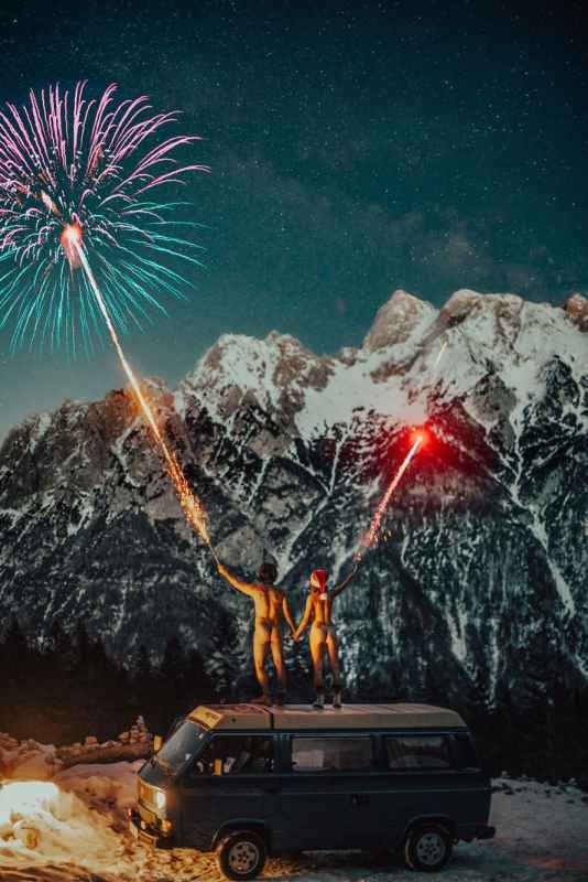 Couple standing on a van roof holding fireworks under a starry sky with snowy mountains.