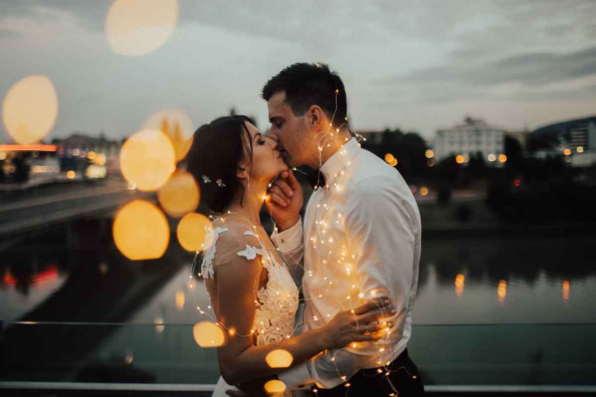 Romantic couple sharing a kiss surrounded by glowing bokeh fairy lights at dusk near a waterfront.