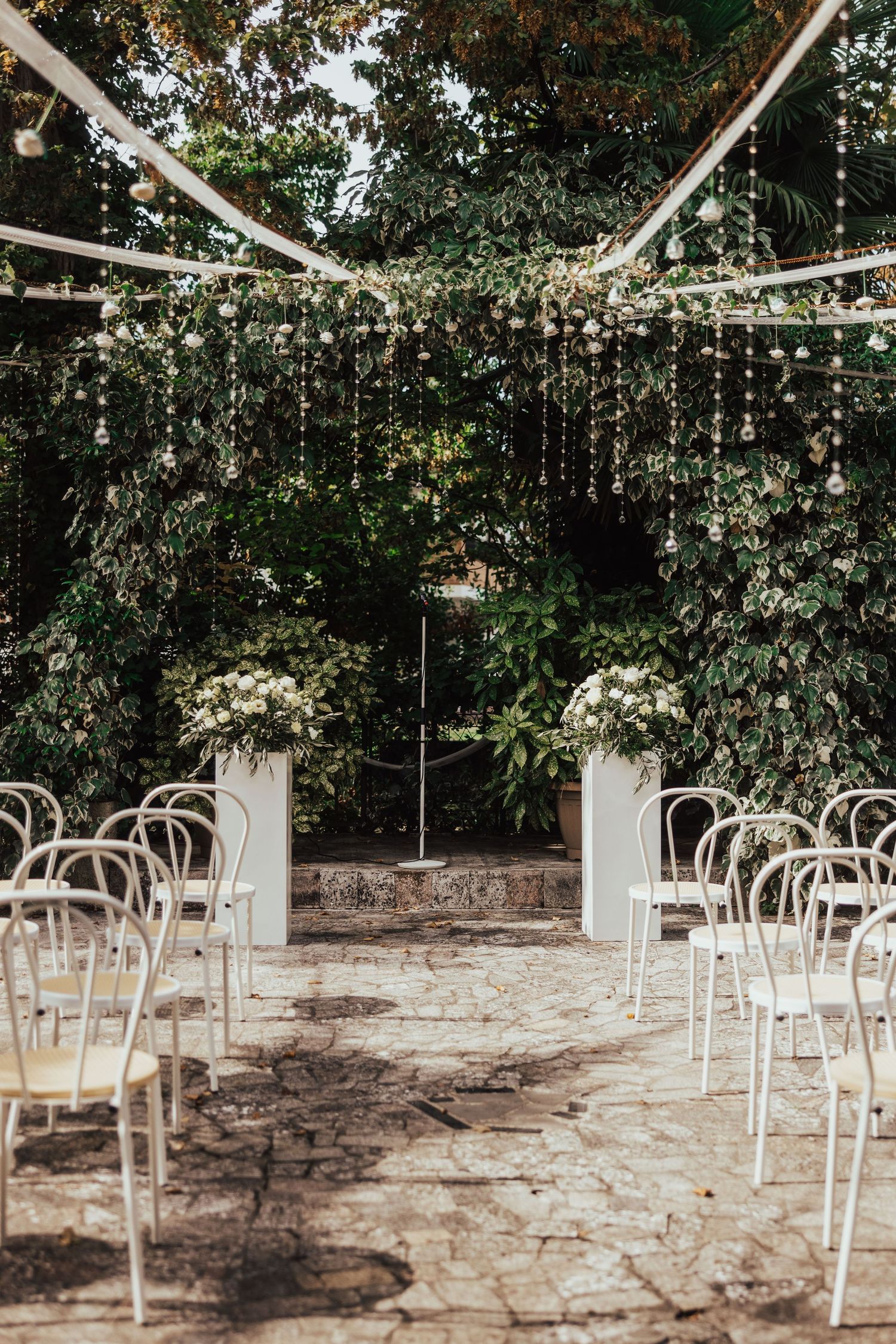 Outdoor wedding ceremony venue with white chairs, floral arch, and hanging crystal decorations in lush garden.