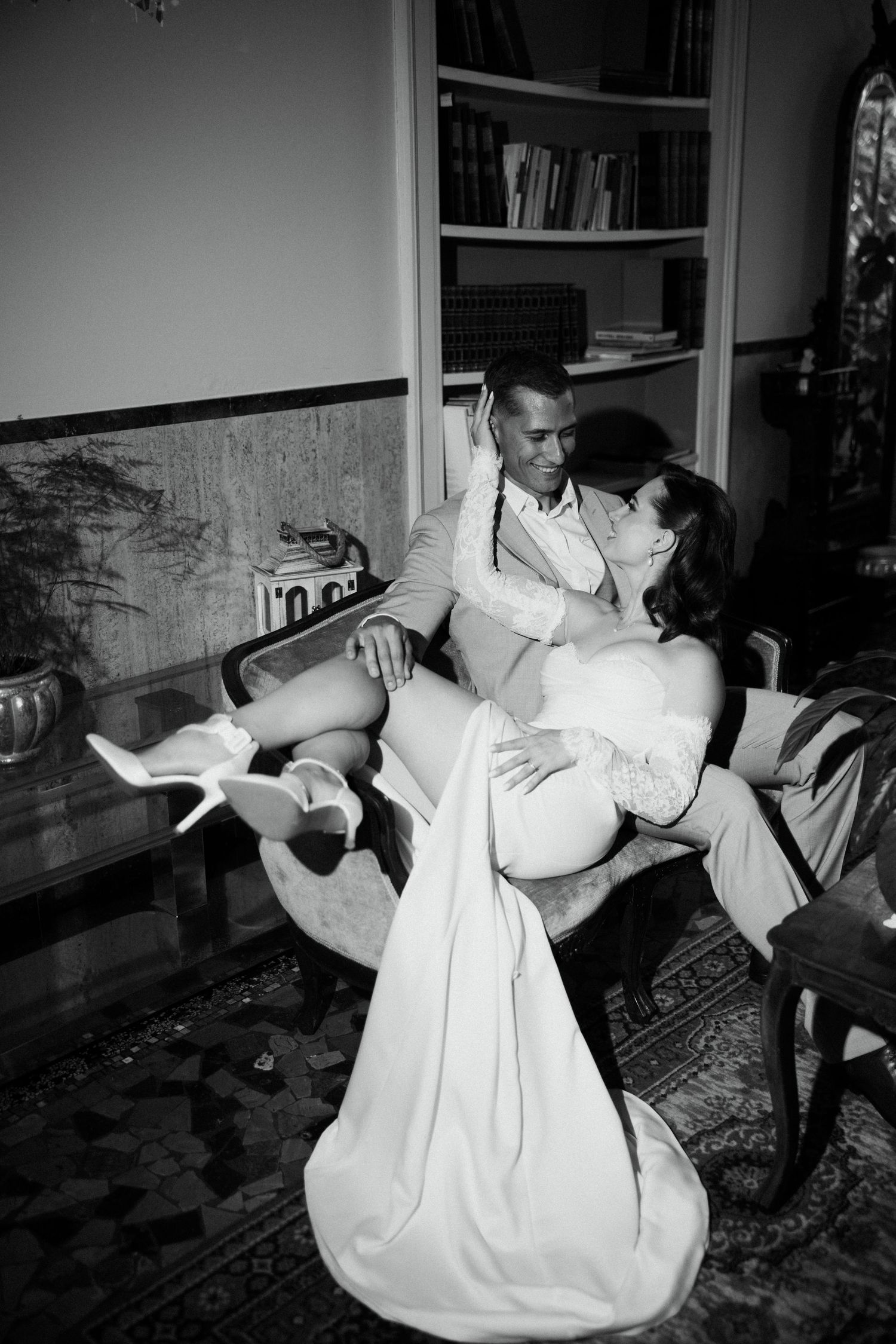 Black and white romantic portrait of bride and groom embracing in vintage interior with bookshelf backdrop.