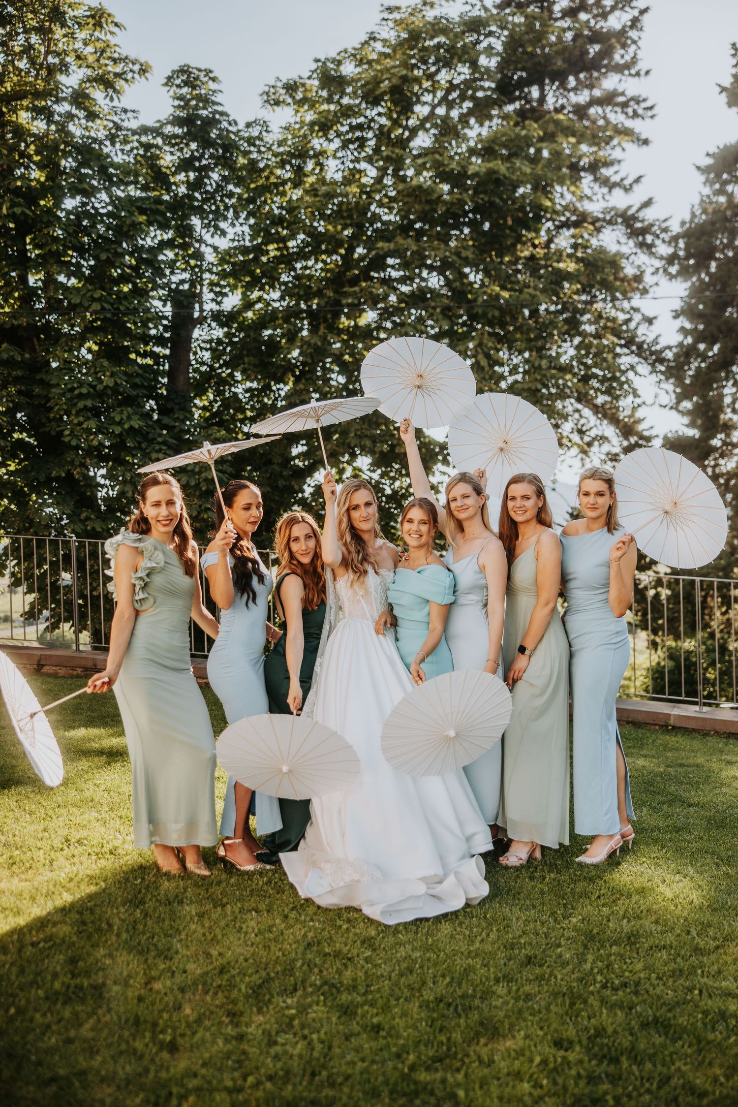 Bride posing with six bridesmaids in sage green dresses holding white parasols outdoors.