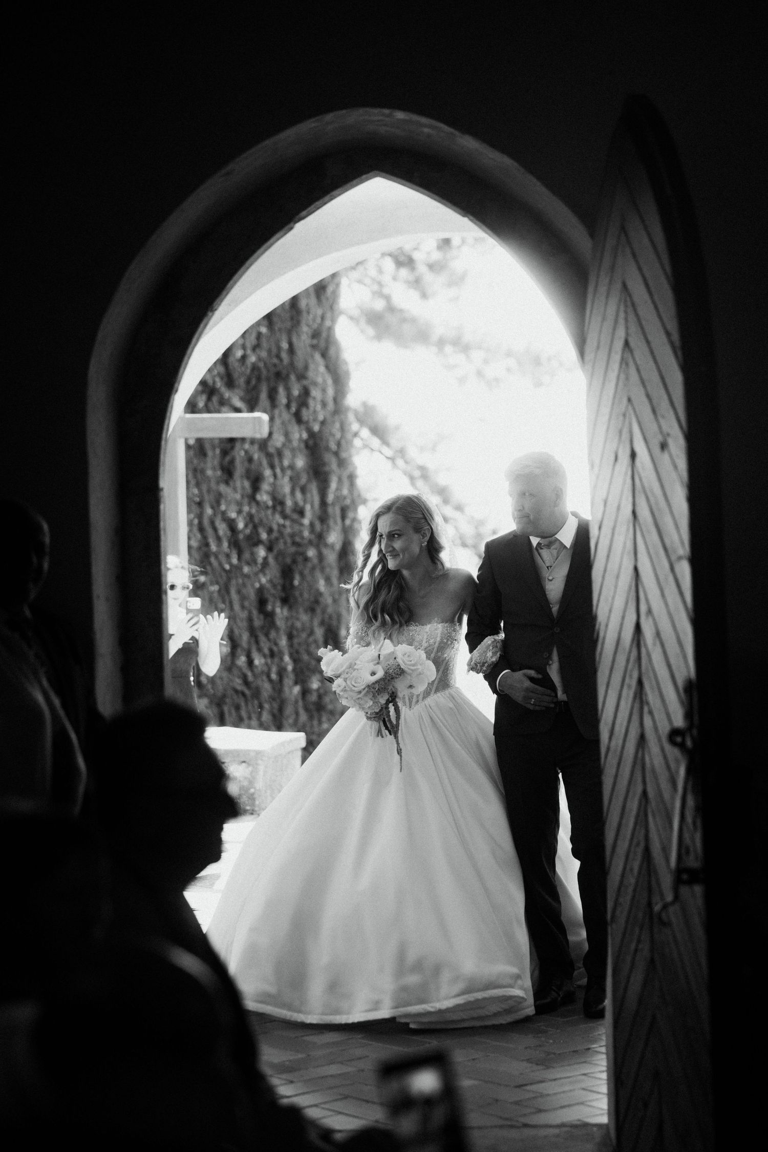 Black and white photo of bride and groom entering church through arched wooden doors.