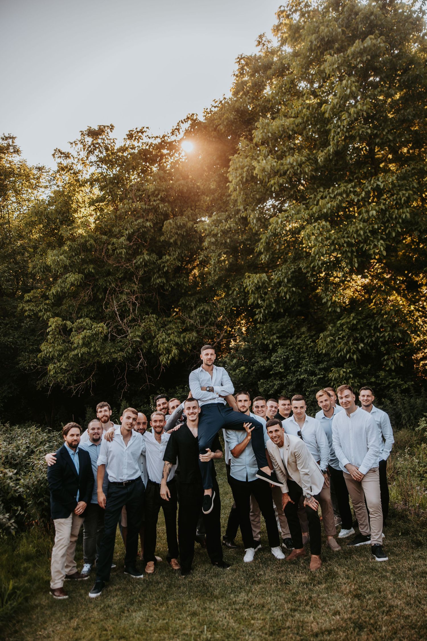 Large group of young men posing outdoors at golden hour, one man lifted on shoulders, surrounded by lush green trees.