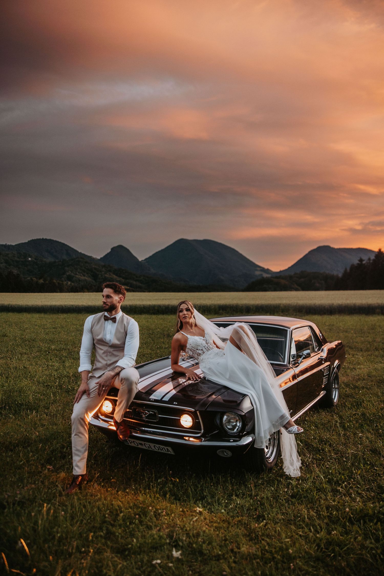 Couple sitting on classic Ford Mustang hood during dramatic sunset.