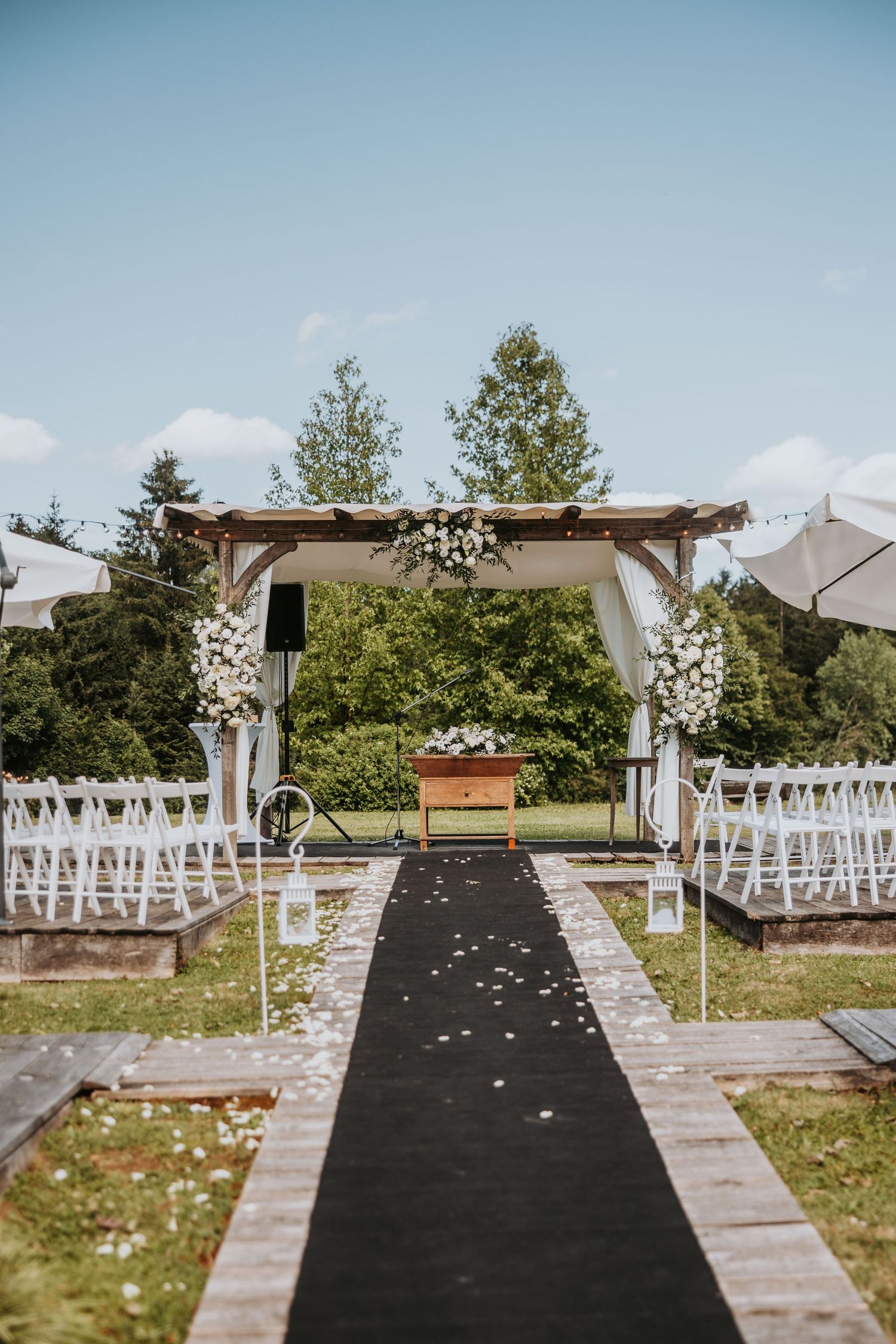 Elegant outdoor wedding ceremony setup with black aisle runner, floral arch, and white chairs on a sunny day.