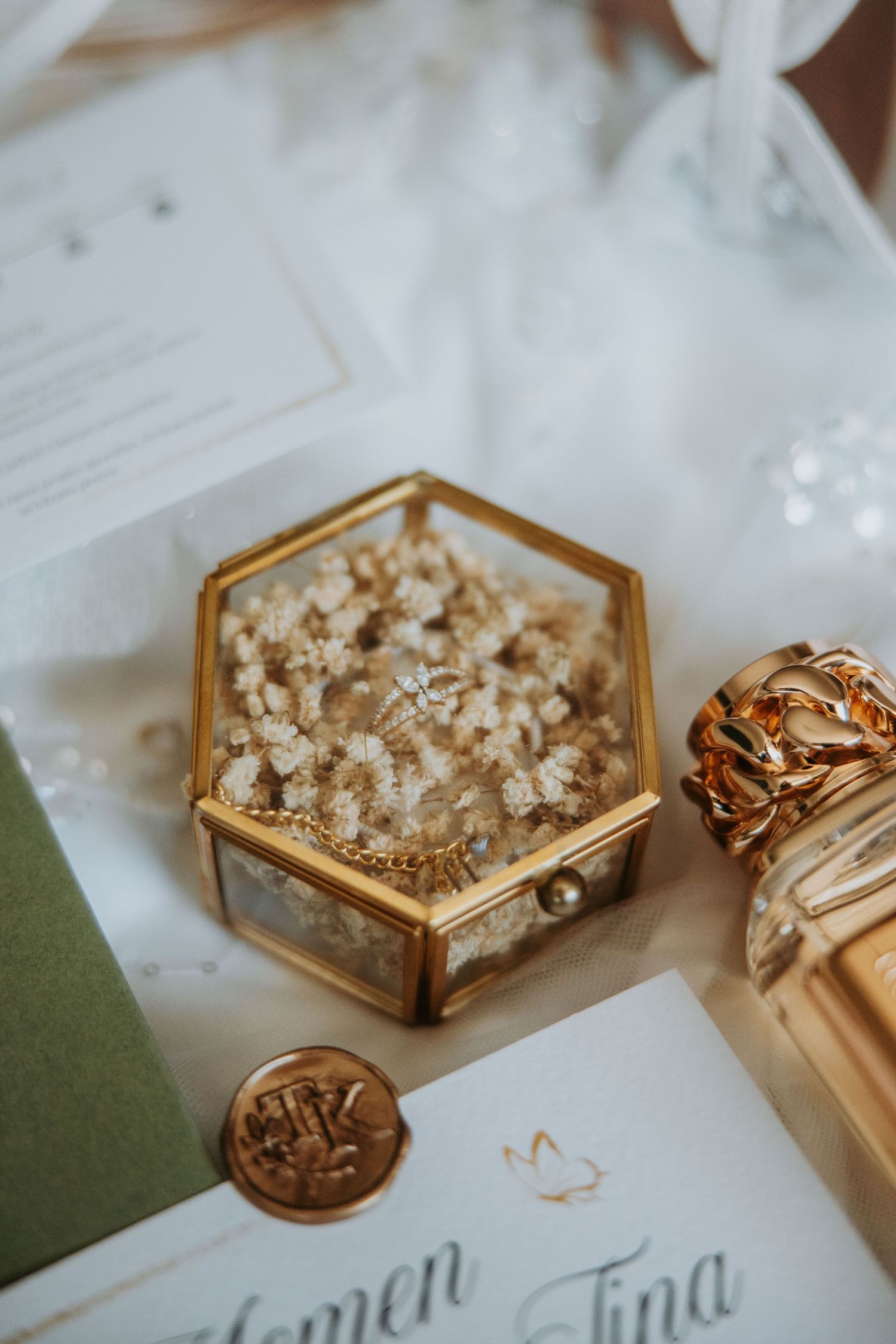 Gold hexagonal glass jewelry box with wedding rings on dried flowers, beside wax seal and elegant wedding stationery.