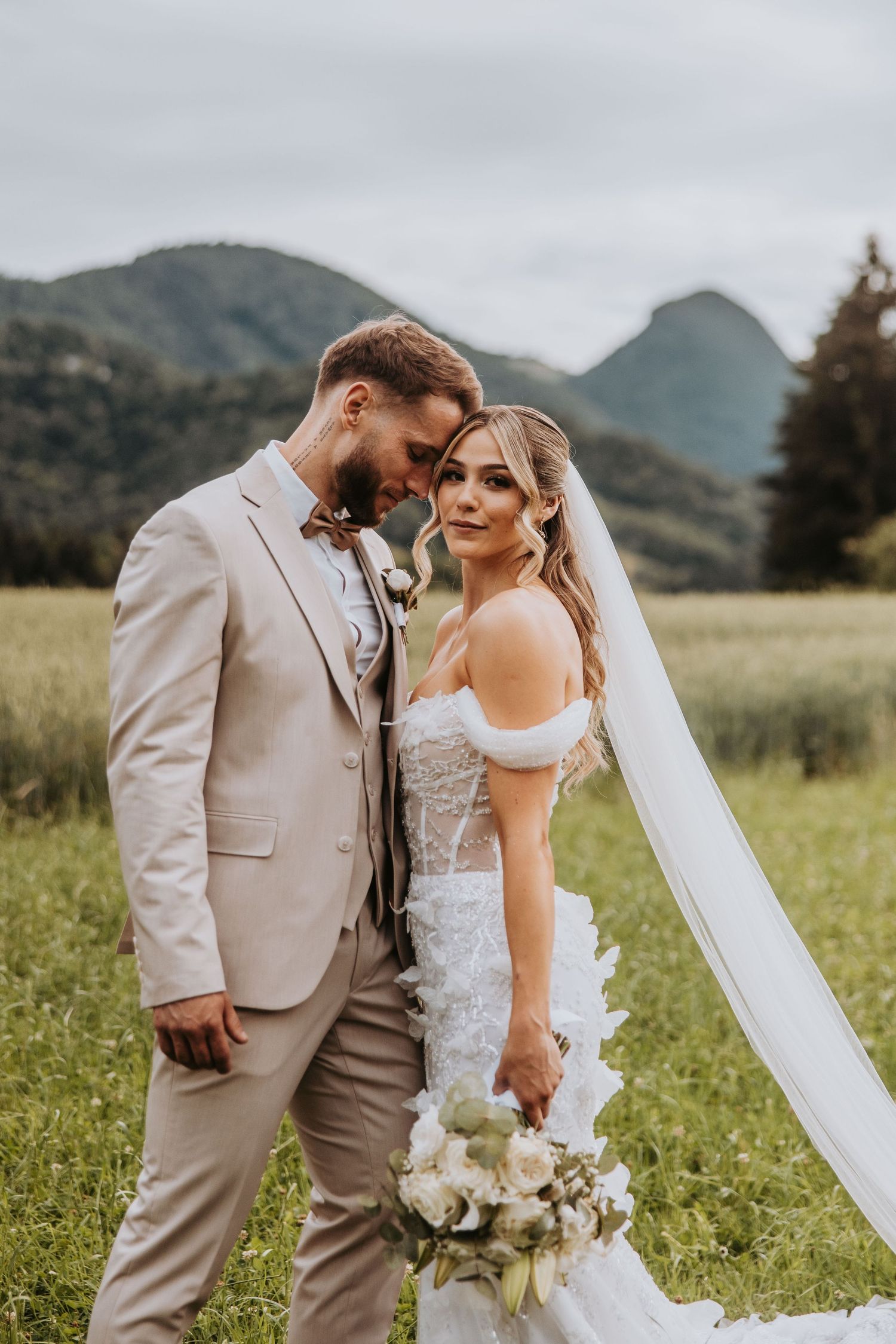 Bride and groom share intimate moment in green meadow with mountain backdrop, bride holds white bouquet.