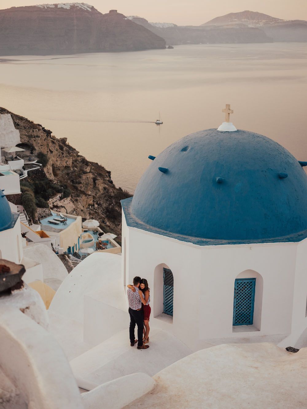 Romantic couple embracing near iconic blue-domed church in Santorini overlooking the caldera at dusk.