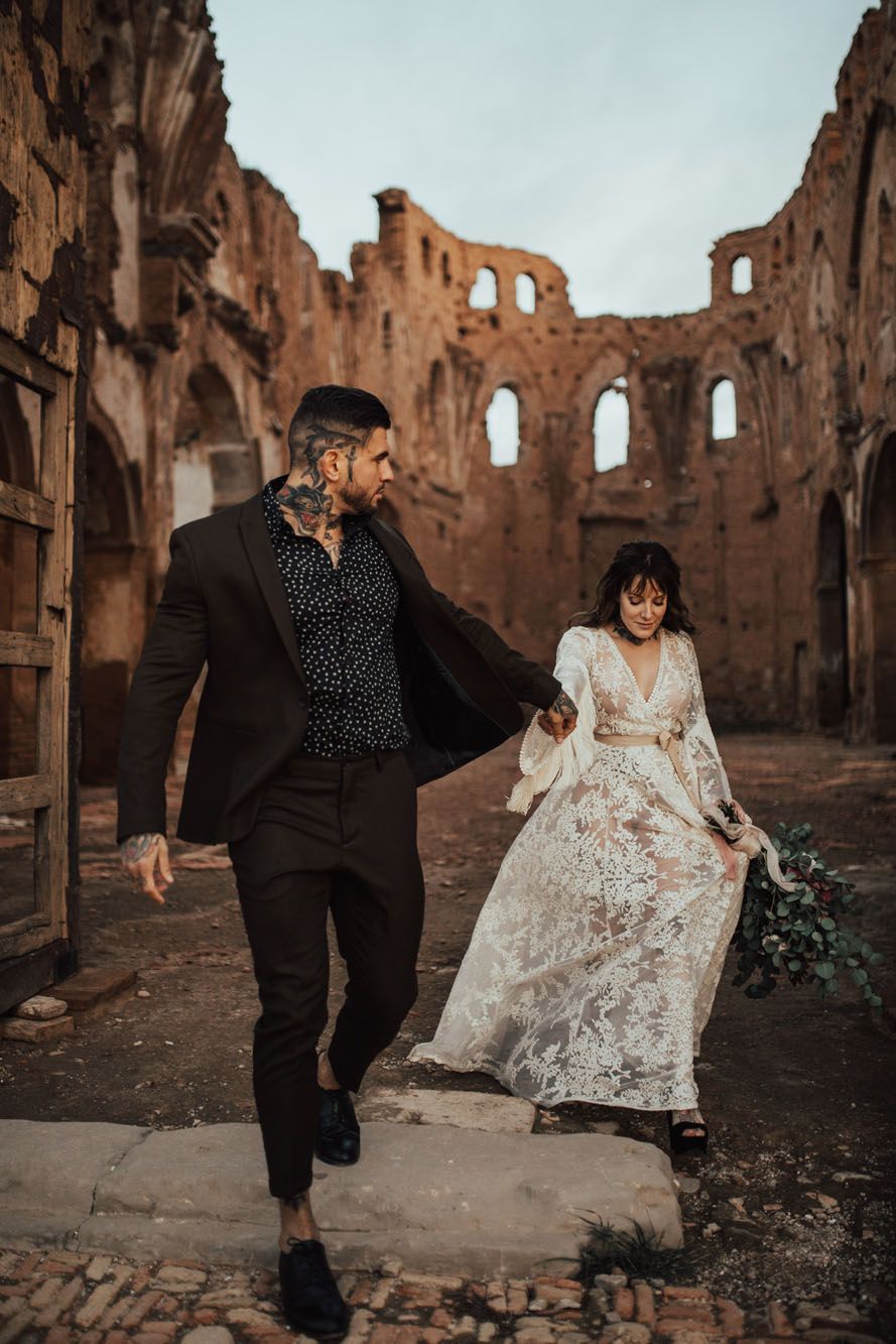 Tattooed groom leading bride through dramatic ancient ruins in lace wedding gown at sunset.