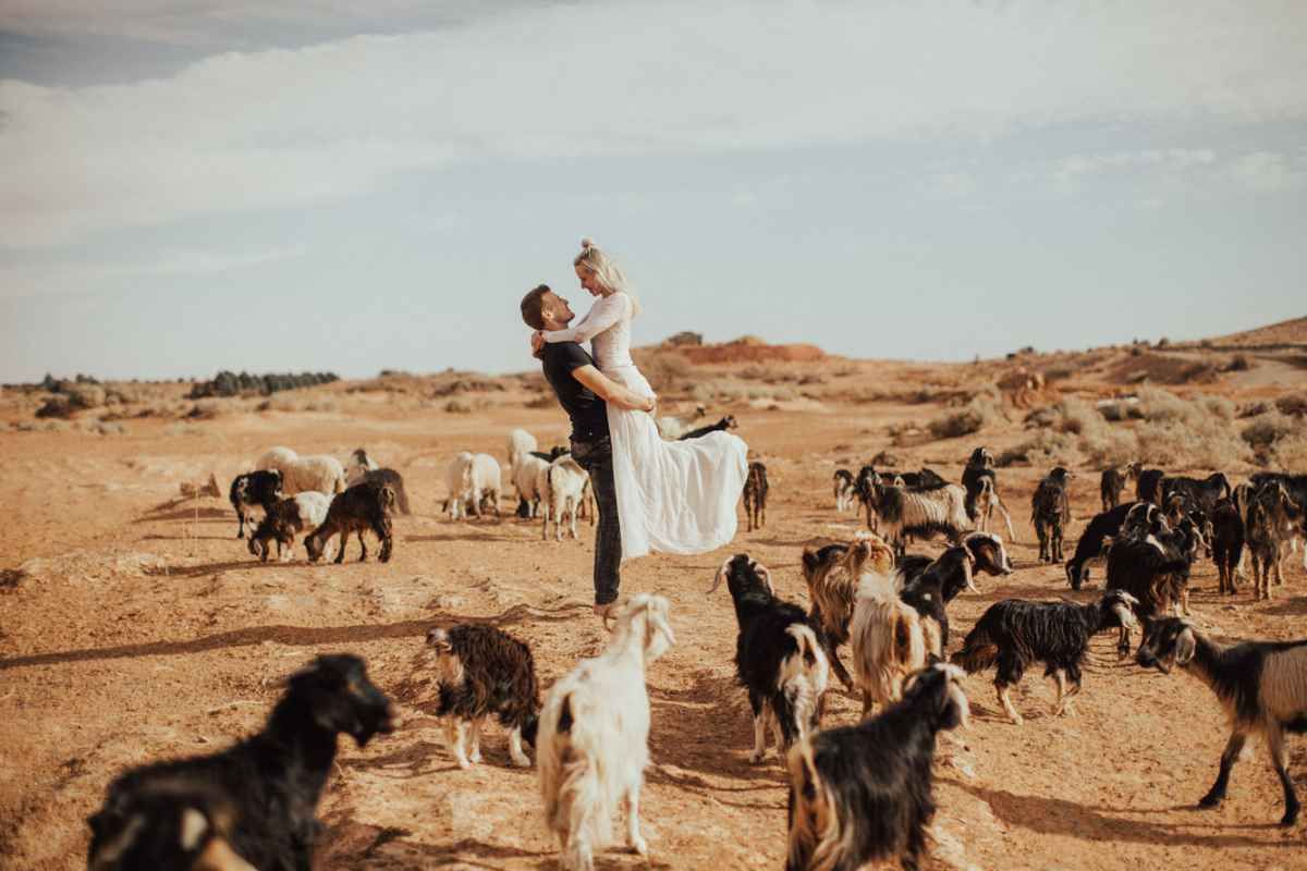 Groom lifting bride in white dress among a herd of goats in a vast desert landscape wedding photo.