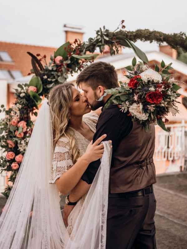 Bride and groom sharing first kiss under floral arch with red and pink roses at wedding.