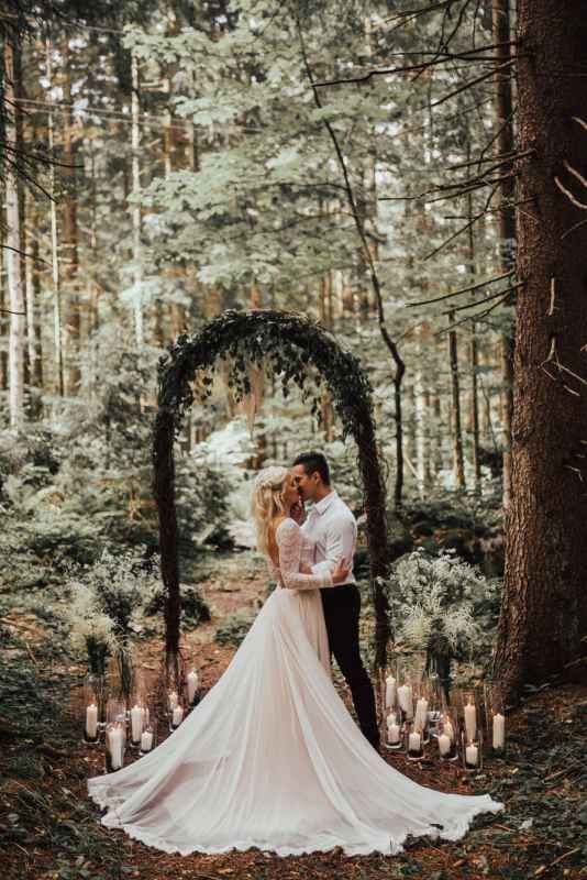 Romantic couple embracing under floral arch with candles in enchanted forest wedding ceremony.
