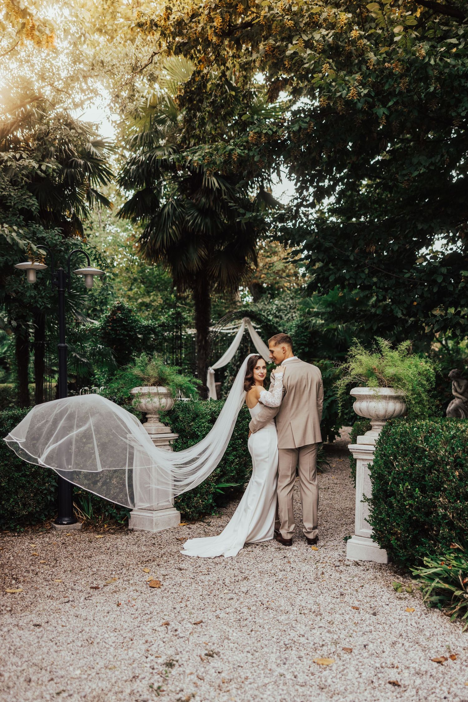 Romantic bride and groom embracing in a sunlit garden with veil flowing in the breeze.