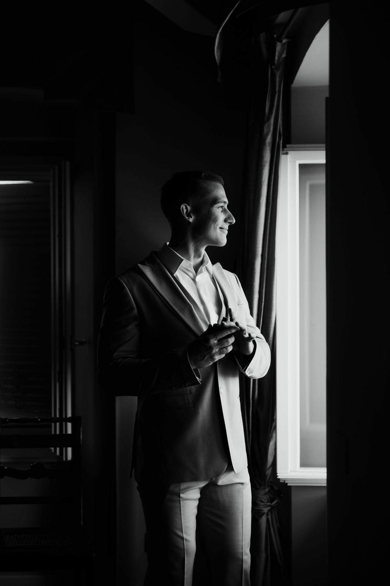 Elegant man in suit standing by window in dramatic black and white portrait photography.